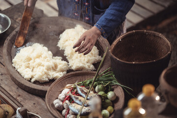 From above, a woman prepares sticky rice next to a platter of grilled fish and fresh vegetables. This shot captures the complete, balanced composition of a traditional Isaan meal.