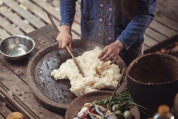 A close-up view shows a woman's hands skillfully using a paddle to mix steaming hot sticky rice. This image highlights the traditional technique and texture of this beloved Thai food.