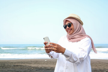 Happy Asian woman using mobile phone chatting or online booking hotel at the beach. People enjoy outdoor lifestyle travel nature the sea with digital gadget device on summer beach holiday vacation.