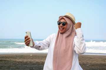 Excited beautiful Asian woman in green sweater and hijab using a mobile phone, celebrating success,...