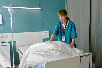 Young adult Caucasian woman nurse making hospital bed in patient room, standing beside medical...