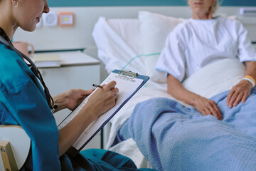 Caucasian female doctor writing on clipboard while sitting beside senior Caucasian woman lying in...