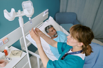 Caucasian young adult female nurse adjusting intravenous drip beside hospital bed with middle aged biracial man lying down receiving medical treatment in clinical setting