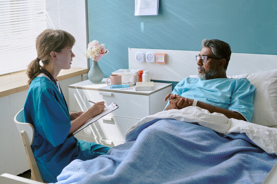 Caucasian young adult female nurse sitting beside hospital bed talking to middle aged biracial man patient lying under blanket holding hands during medical consultation
