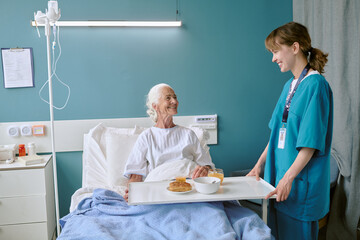 Senior Caucasian woman sitting in hospital bed smiling at young adult Caucasian female nurse...