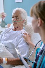 Senior Caucasian woman lying in hospital bed wearing oxygen tube, receiving support from young...