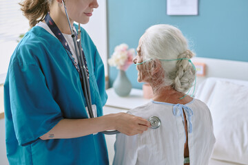 Caucasian female healthcare worker examining senior Caucasian woman with stethoscope in medical setting, senior woman sitting on hospital bed with oxygen tube, healthcare professional standing
