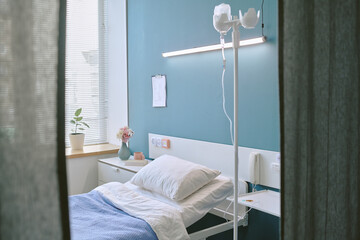 Empty hospital bed positioned near window with medical equipment and clipboard on wall, showing...