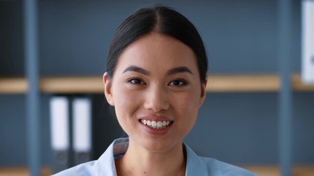A young Asian woman, dressed professionally, smiles warmly at the camera in a modern office environment. She appears confident and engaged while working in a collaborative space.