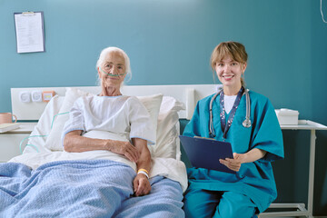 Obraz premium Senior Caucasian woman lying in hospital bed with oxygen tube, smiling young adult Caucasian female nurse sitting beside bed holding clipboard, both looking at camera