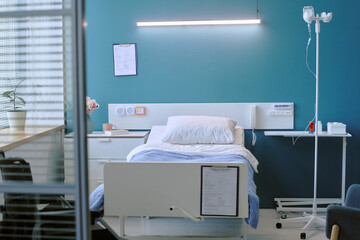 Empty hospital bed standing in modern medical room with clipboard on wall, IV stand nearby, medical...