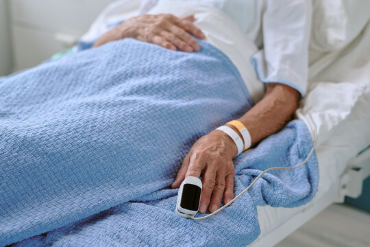 Senior woman lying in hospital bed, resting with hand visible wearing medical wristbands and pulse oximeter sensor attached to finger covered by blue blanket