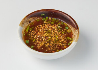 Bowl of flavorful dipping sauce with rich brown base, garnished with chopped green onions and sesame seeds, served in white ceramic bowl with rustic brown rim