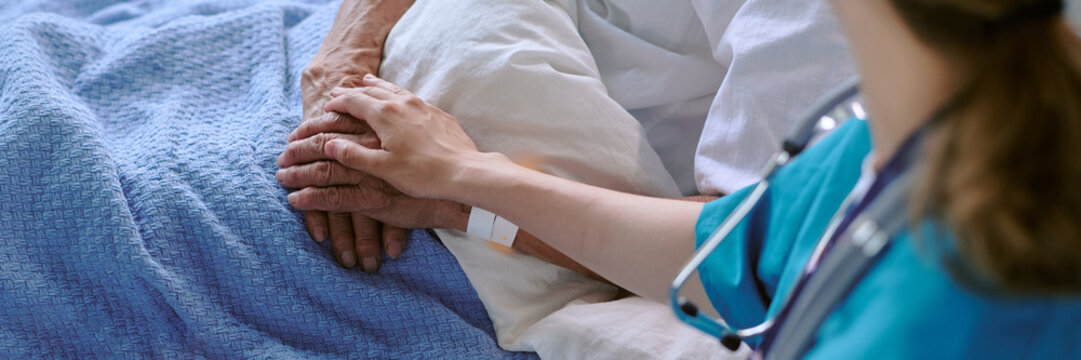 Senior patient lying in hospital bed receiving comforting touch from young female nurse, hands gently clasped together, medical care setting visible - Powered by Adobe