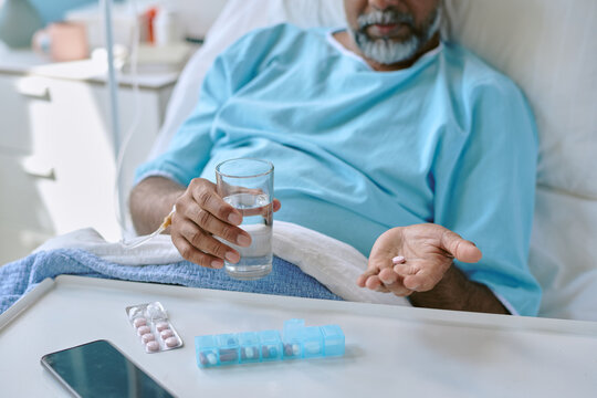 Middle aged biracial man lying in hospital bed holding glass of water in one hand and several pills in other hand, preparing to take medication, pill organizer and smartphone on table