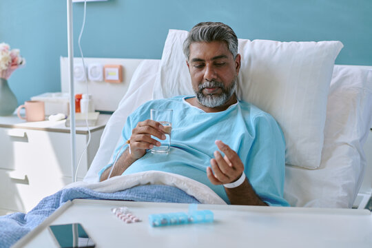 Middle aged biracial man lying in hospital bed holding glass of water and examining medication tablet, wearing patient wristband, medical equipment and pills visible on bedside table - Powered by Adobe