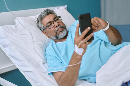 Middle aged biracial man lying in hospital bed using smartphone, wearing medical gown with IV catheter in hand, looking at device screen while resting in patient room