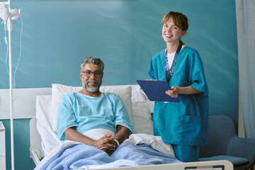 Middle aged biracial man sitting in hospital bed smiling while young adult biracial female nurse...