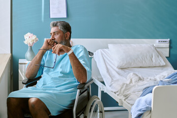 Middle aged biracial man with disability sitting in wheelchair beside hospital bed holding...
