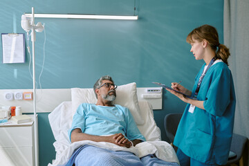Middle aged biracial man lying in hospital bed listening to young adult biracial female nurse...