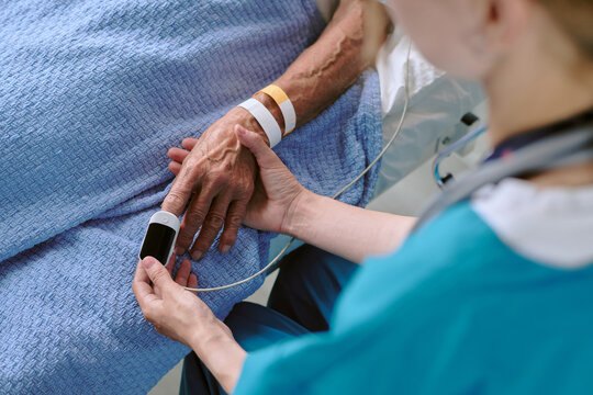 Middle aged biracial man lying in hospital bed receiving care from Caucasian female nurse checking oxygen saturation with pulse oximeter on finger, close up of hands