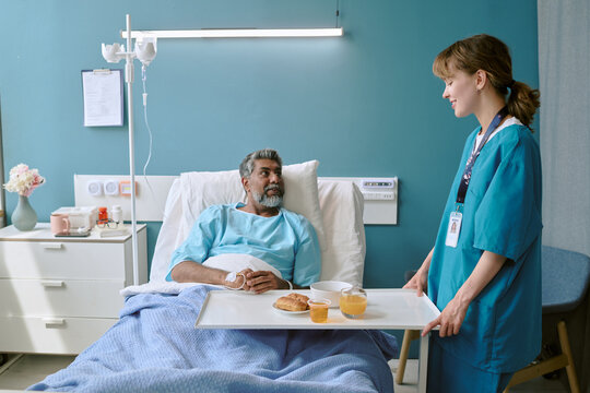 Caucasianl female nurse serving breakfast tray to middle aged biracial man lying in hospital bed, patient looking at nurse, nurse standing beside bed holding tray with food and drinks