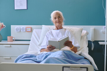 Portrait of senior Caucasian woman sitting in hospital bed reading book, smiling gently while...