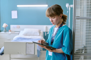 Young adult Caucasian woman nurse standing in hospital room using digital tablet, focusing on patient records, medical professional working in healthcare environment
