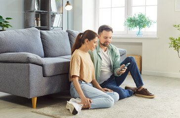 Young couple sitting on floor of their cozy home using smart phone together, browsing social media,...