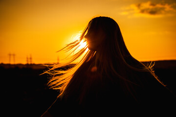 Silhouette of a teenage girl with long hair in the wind against a golden sunset sky, evoking a...