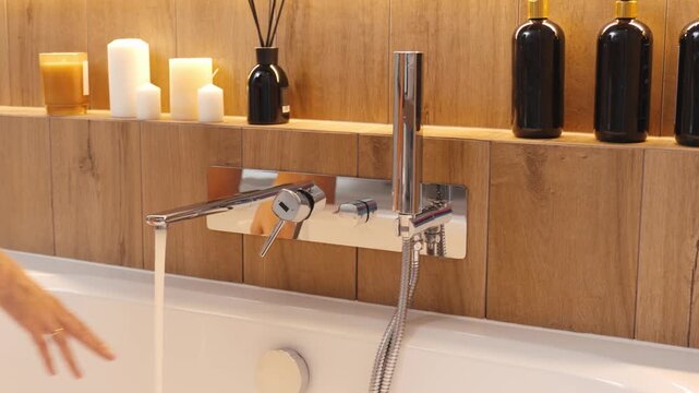 Close-up of person hand turning chrome bathtub faucet, with water flowing into white tub. Woman adjusting water temperature in bathroom