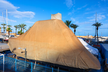 Covered yacht in dry dock, secured under a large protective beige tarp, surrounded by palm trees and moored boats under a clear blue sky. Maritime maintenance scene in a sunny Mediterranean marina