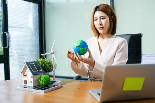 Confident woman holding globe in eco office, working on clean energy solutions with wind turbine, solar house, and green business strategy.