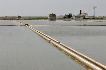 Concrete canals intersecting in salt evaporation ponds with industrial buildings in the background,...