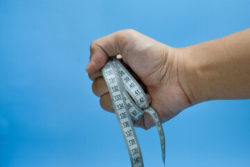 A human hand tightly gripping a white measuring tape with black numbers, set against a clean blue...
