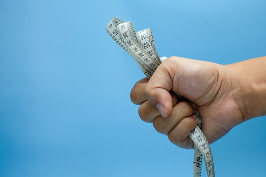 A human hand clenched into a fist, gripping a soft measuring tape tightly against a blue background. Ideal for health campaigns, fitness goals, discipline, health tracking or wellness advertisements - Powered by Adobe