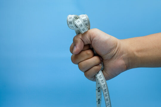 A human hand clenched into a fist, gripping a soft measuring tape tightly against a blue background. Ideal for health campaigns, fitness goals, discipline, health tracking or wellness advertisements