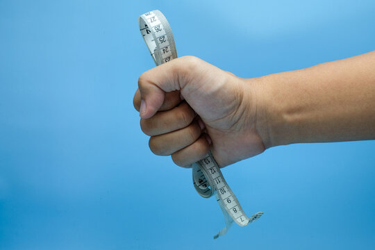 A human hand clenched into a fist, gripping a soft measuring tape tightly against a blue background. Ideal for health campaigns, fitness goals, discipline, health tracking or wellness advertisements