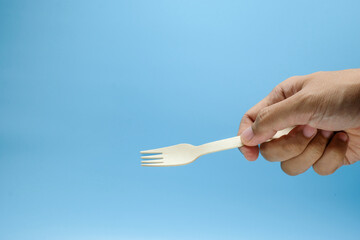 A hand is holding a wooden disposable fork with a clean blue background. Suitable for eco-friendly...