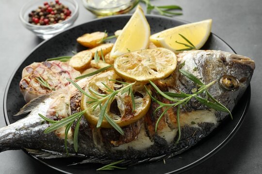 Tasty roasted dorado fish with potatoes, rosemary and lemon on grey table, closeup. Homemade seafood dish