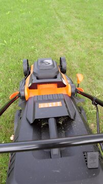 POV gardener cuts the green grass on lawn with an orange electric lawnmower. Vertical video.