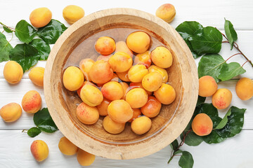 Fresh ripe apricots in bowl with water and green leaves on white wooden table, flat lay