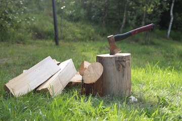 Metal axe, chopped firewood and wooden log on green grass outdoors