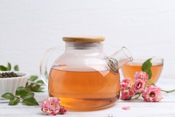 Tasty rose tea in glass teapot, flowers and leaves on white wooden table, closeup