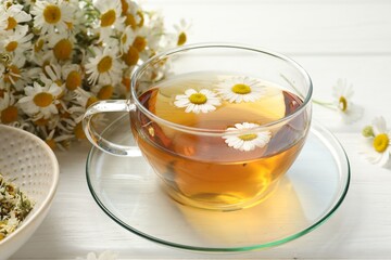 Aromatic tea in glass cup and chamomile flowers on white wooden table, closeup