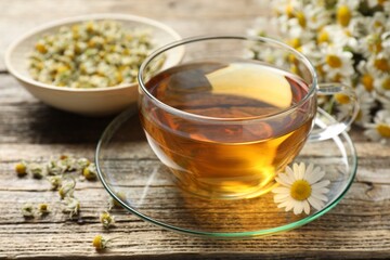 Aromatic tea in glass cup and chamomile flowers on wooden table, closeup