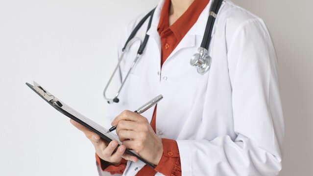Unknown woman doctor wearing stethoscope and white coat over brick red blouse is standing and writing on a clipboard, representing medical expertise and patient care. Medicine and health care concept