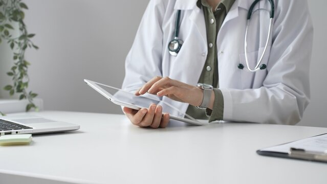 Doctor woman in lab coat and stethoscope using digital tablet at desk in medical office, accessing online patient information. Medicine and health care concept