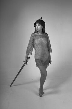 Full length black and white portrait of young warrior woman in chainmail and original medieval Asian helmet holding sword on grey background