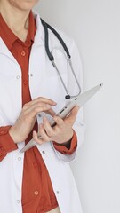 Unknown professional woman doctor, wearing white coat over a brick-red blouse and a stethoscope, is...